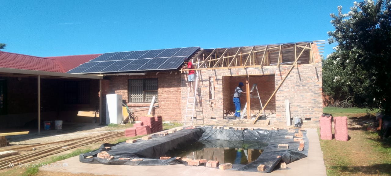 Completed red tile roof on brick extension, solar panels on adjacent building, workers below, blue sky