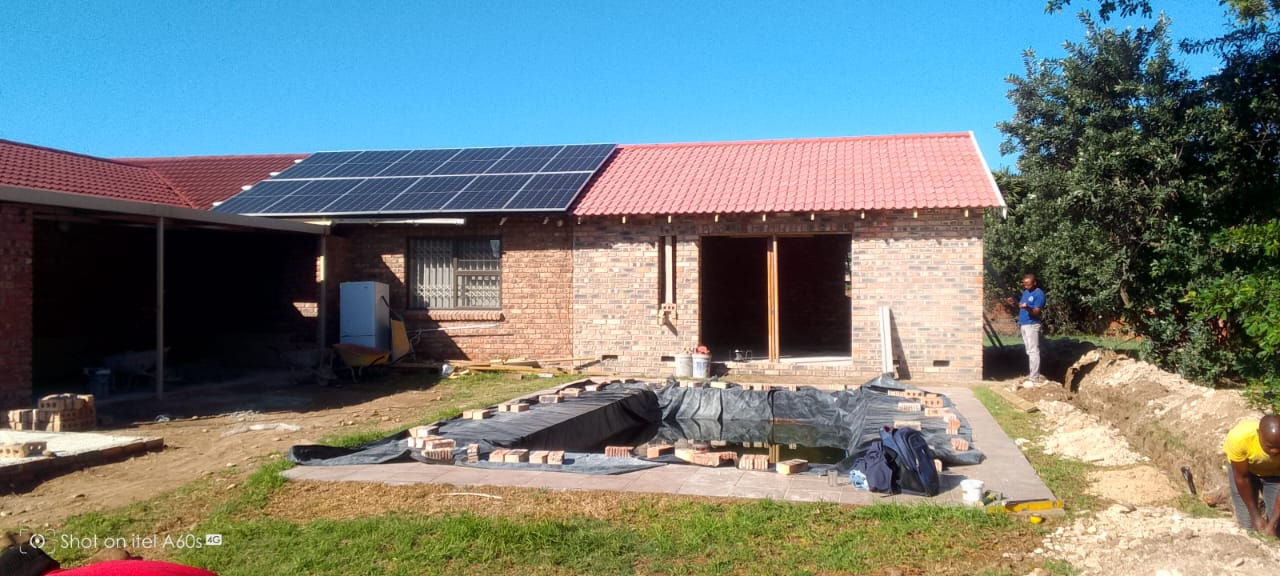 Two workers on red tile roof actively tiling, pool construction visible below, solar panels left